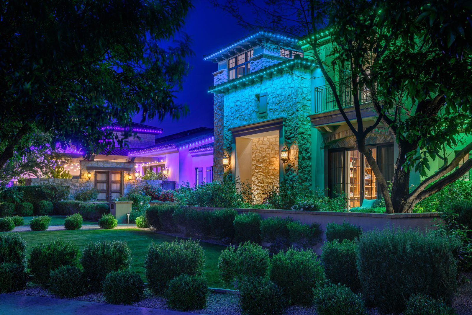 Teal and purple permanent roofline lighting on a stone estate with landscape lighting in the front yard