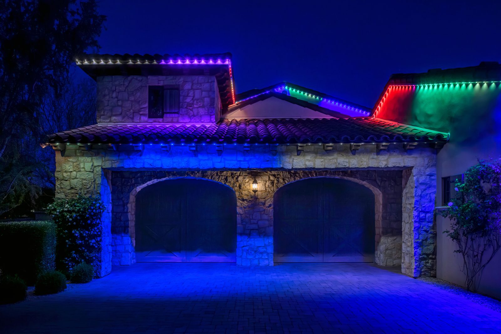 Multicolor permanent roofline lighting over a stone garage with tile roof at night