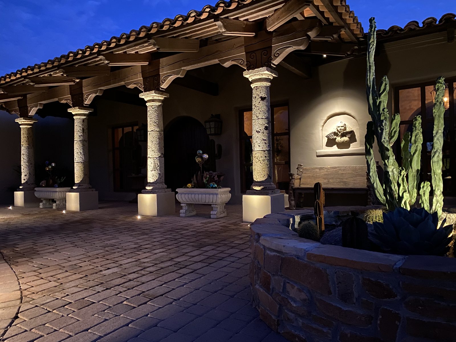 Covered patio with stone columns lit by warm downlights and uplit cactus