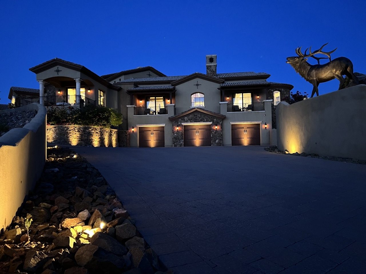 Large estate at dusk with lit garage doors, driveway walls and an elk statue